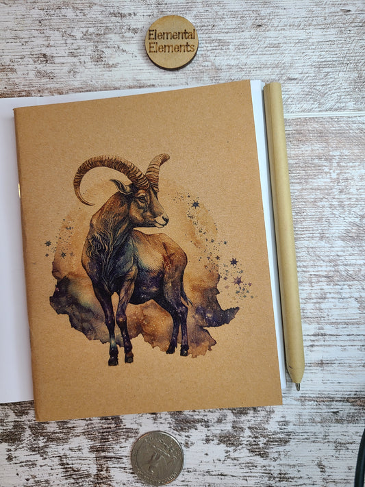 Capricorn Zodiac full bodied ram in browns and teal with teal, blue, and brown galaxy background. Journal is sitting on top of an open journal showing blank pages. An eco-friendly pen lies next to the Zodiac journal.