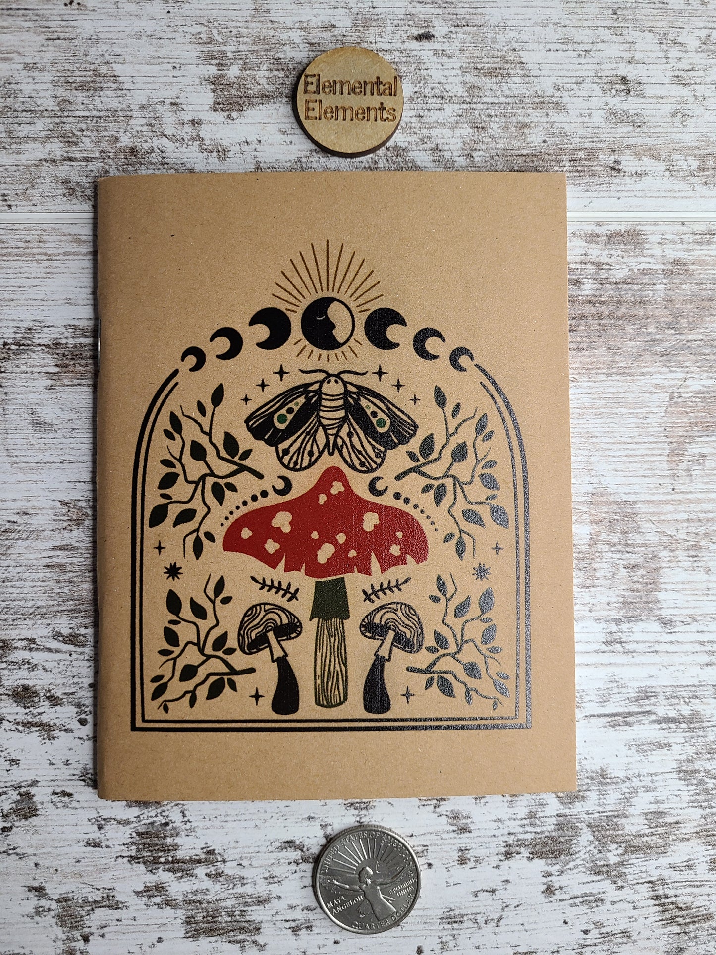 Journal with one red mushroom in the center and other mushrooms, a moth, leaves, moon, and sun surround in black or muted colors.