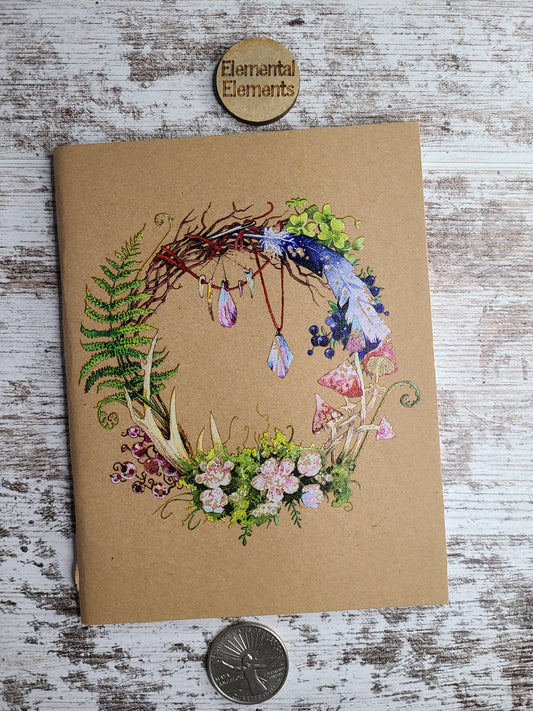 Journal with unique art depicting fern, bones, crystals, berries, flowers, fungi, red string with a needle, and twigs made into a wreathe.