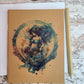 Aquarius zodiac astrology is a human with dark blue and green long flowing hair, blue two-piece clothing, gold accessories in their hair and bottoms. There is a gold detailed u-shaped piece below Aquarius that Aquarius is holding and looking down at, and it is all within a watercolor circle made up of blues, greens, and purple.   Journal is sitting on top of an open journal showing blank pages. An eco-friendly pen lies next to the Zodiac journal.