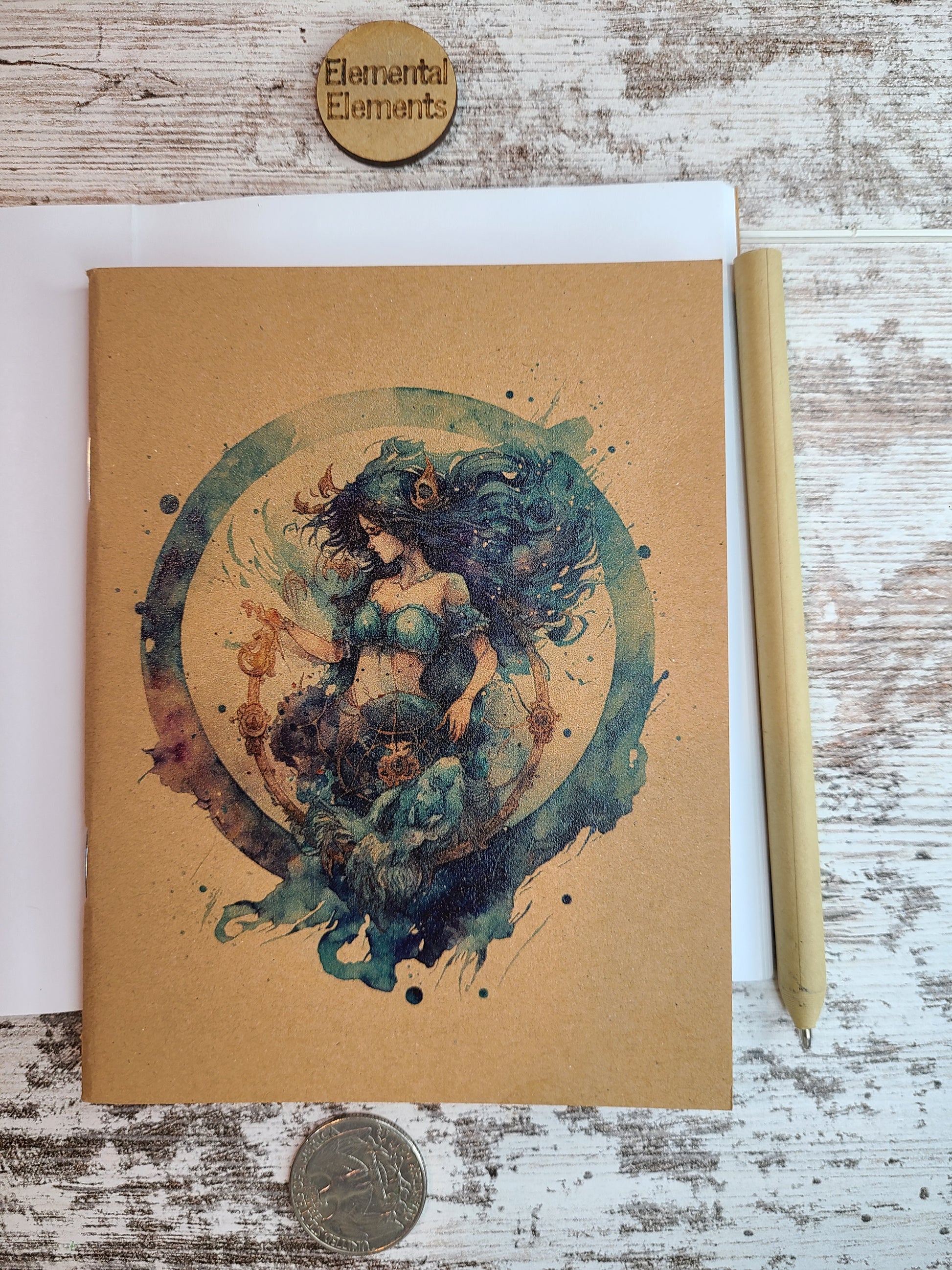 Aquarius zodiac astrology is a human with dark blue and green long flowing hair, blue two-piece clothing, gold accessories in their hair and bottoms. There is a gold detailed u-shaped piece below Aquarius that Aquarius is holding and looking down at, and it is all within a watercolor circle made up of blues, greens, and purple.   Journal is sitting on top of an open journal showing blank pages. An eco-friendly pen lies next to the Zodiac journal.