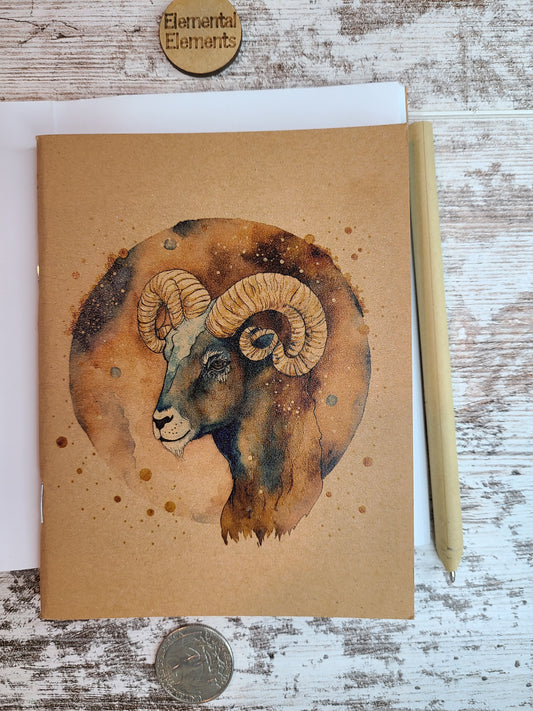 Aries Zodiac journal shows the side view of the head of a ram in shades of browns and blues. Aries has a brown and blue watercolor circle background. The Aries astrology journal is sitting on top of an open journal showing blank pages. An eco-friendly pen lies next to the Zodiac journal.