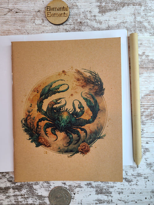 Cancer zodiac astrology journal shows a water color depiction of a large blue and brown crab within a brown, tan, and blue circle that has reef and floral elements surround. Journal is sitting on top of an open journal showing blank pages. An eco-friendly pen lies next to the Zodiac journal.